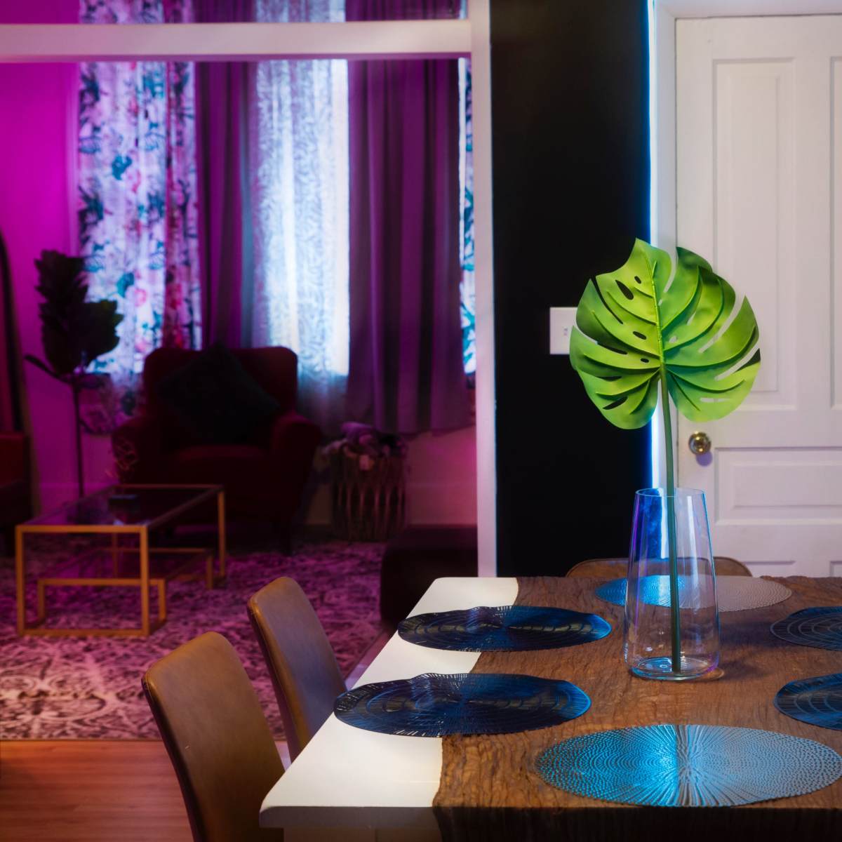 A dining table with blue plates and a vase containing a large green leaf sits in a room accented by purple lighting and floral curtains.