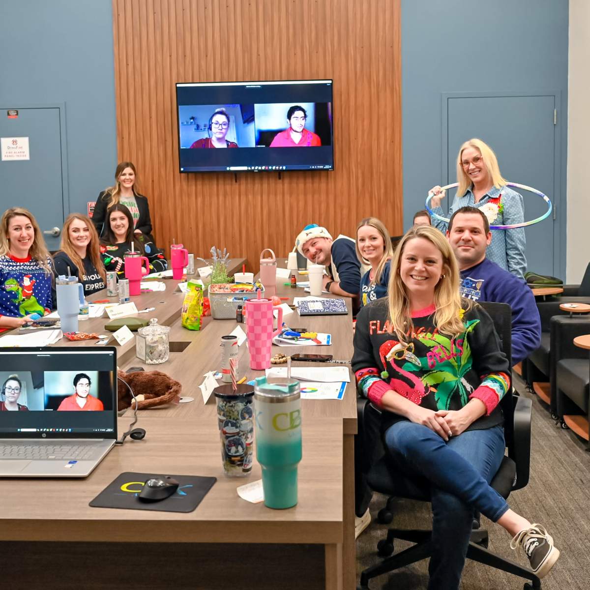 A group of twelve people is gathered around a conference table in a well-lit meeting room, some wearing festive sweaters, while others appear on a video call displayed on a large screen.