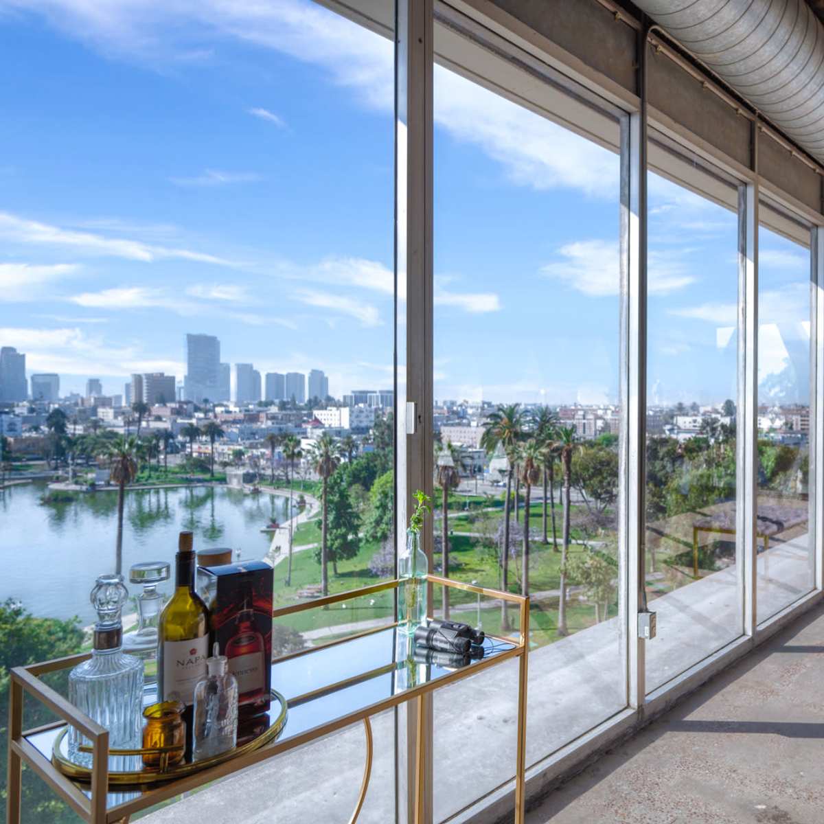 A spacious interior with large windows overlooking a cityscape and a body of water, featuring a table with various bottles and glasses.