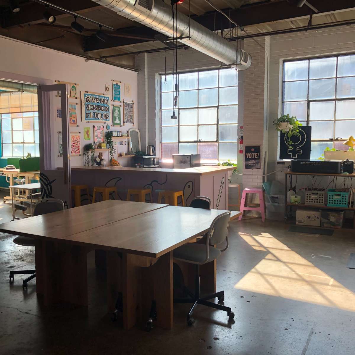 A spacious, well-lit communal workspace with wooden tables, large windows, and colorful decor.