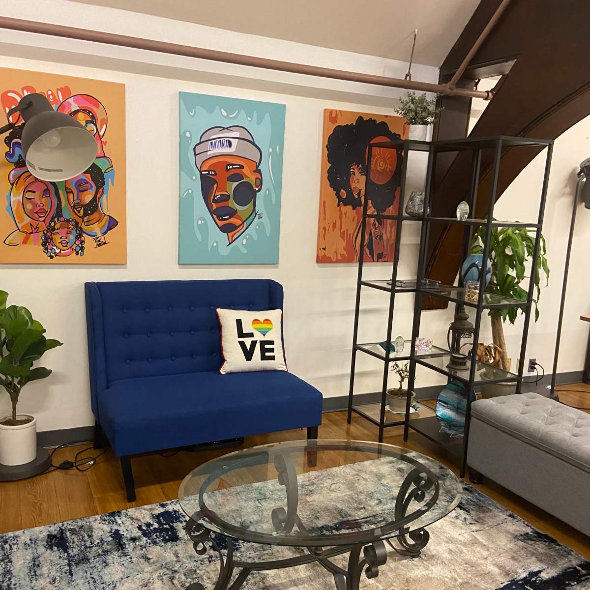 A blue couch with a "LOVE" pillow, a glass coffee table, and colorful artwork displayed on the walls in a modern living room setting.
