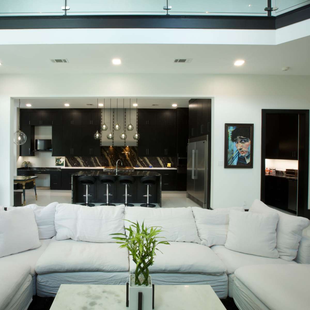 A modern open-concept living space with a large white sectional sofa in the foreground, leading to a kitchen area featuring dark cabinetry and pendant lighting.