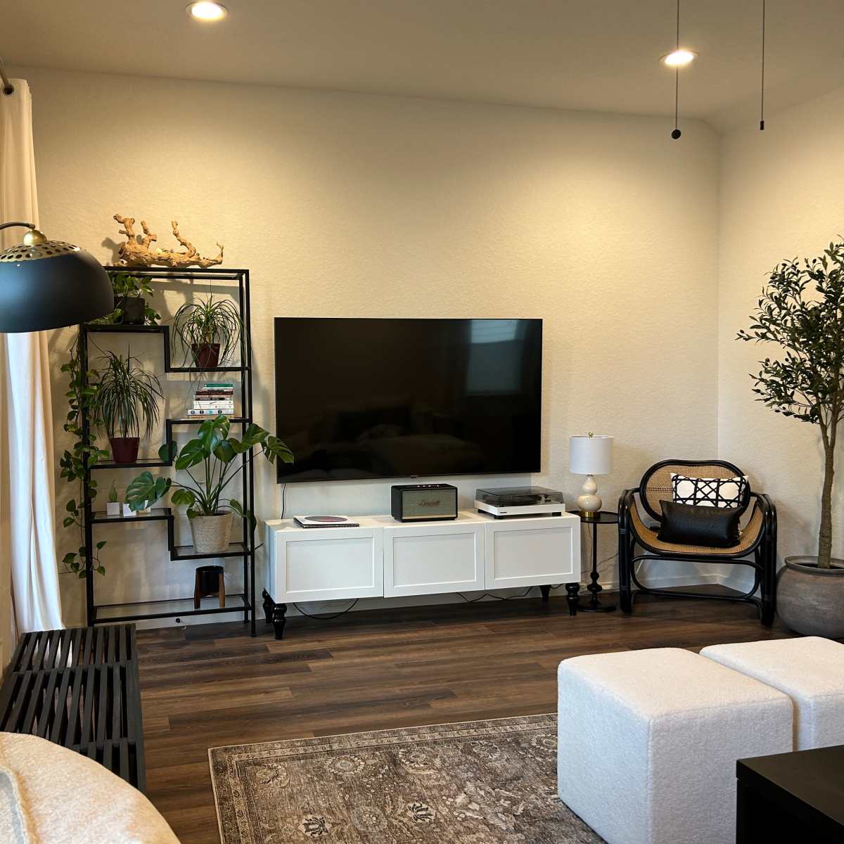 A modern living room features a large TV mounted on a beige wall, surrounded by plants, a bookshelf, and a white cabinet with a sleek black lamp and a wicker chair.