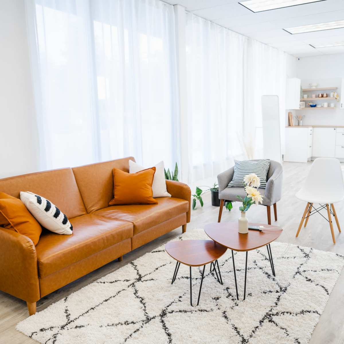 A bright and airy room features a brown leather sofa, two coffee tables, a gray armchair, and a modern kitchen area in the background.