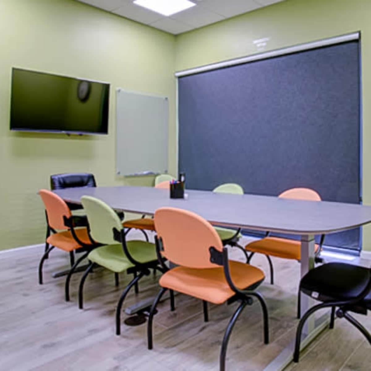 A meeting room features a large table surrounded by chairs in orange and black, with a TV mounted on the wall and a clock above the door.