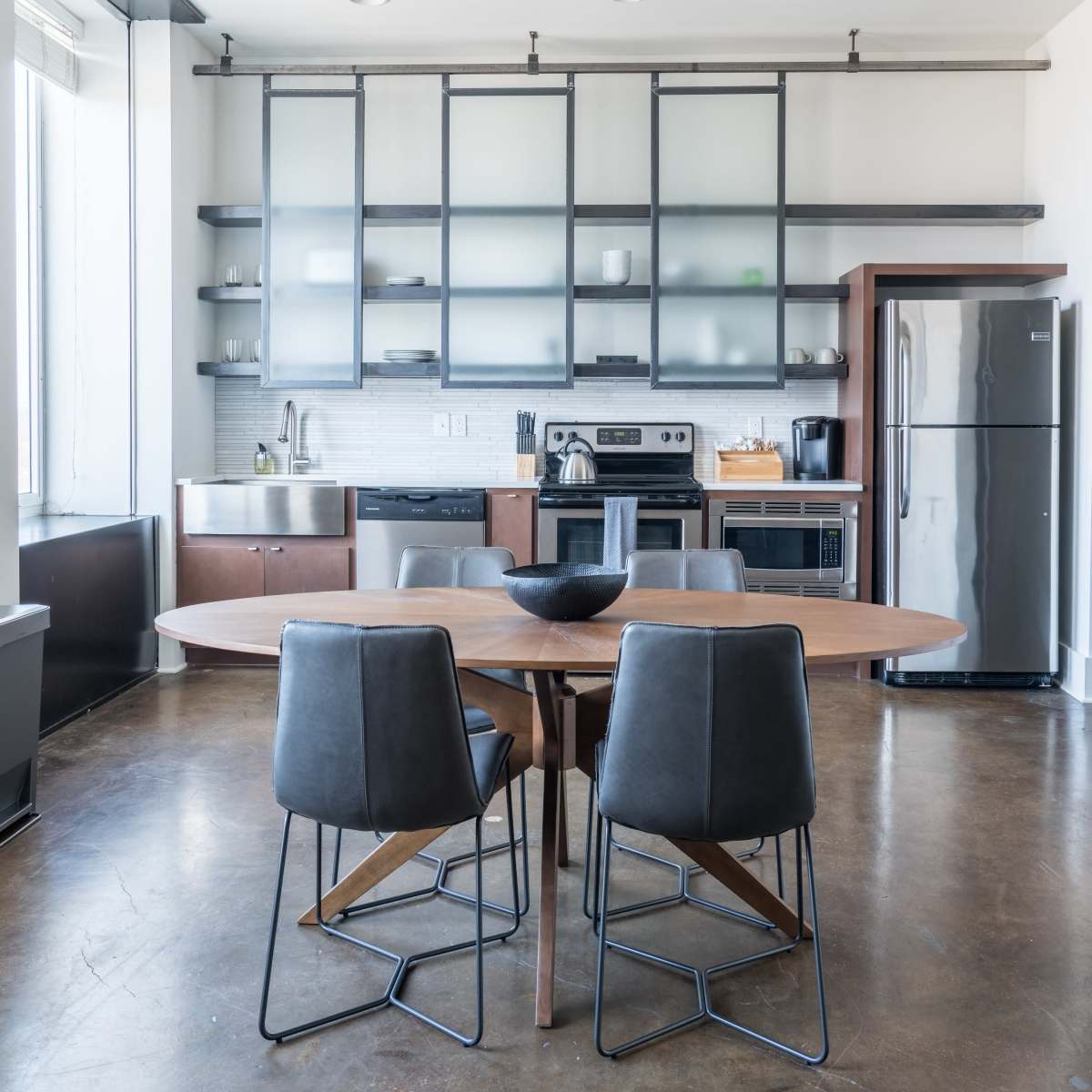 A modern kitchen with a large round wooden table and four black metal chairs, along with stainless steel appliances and glass-front cabinetry.