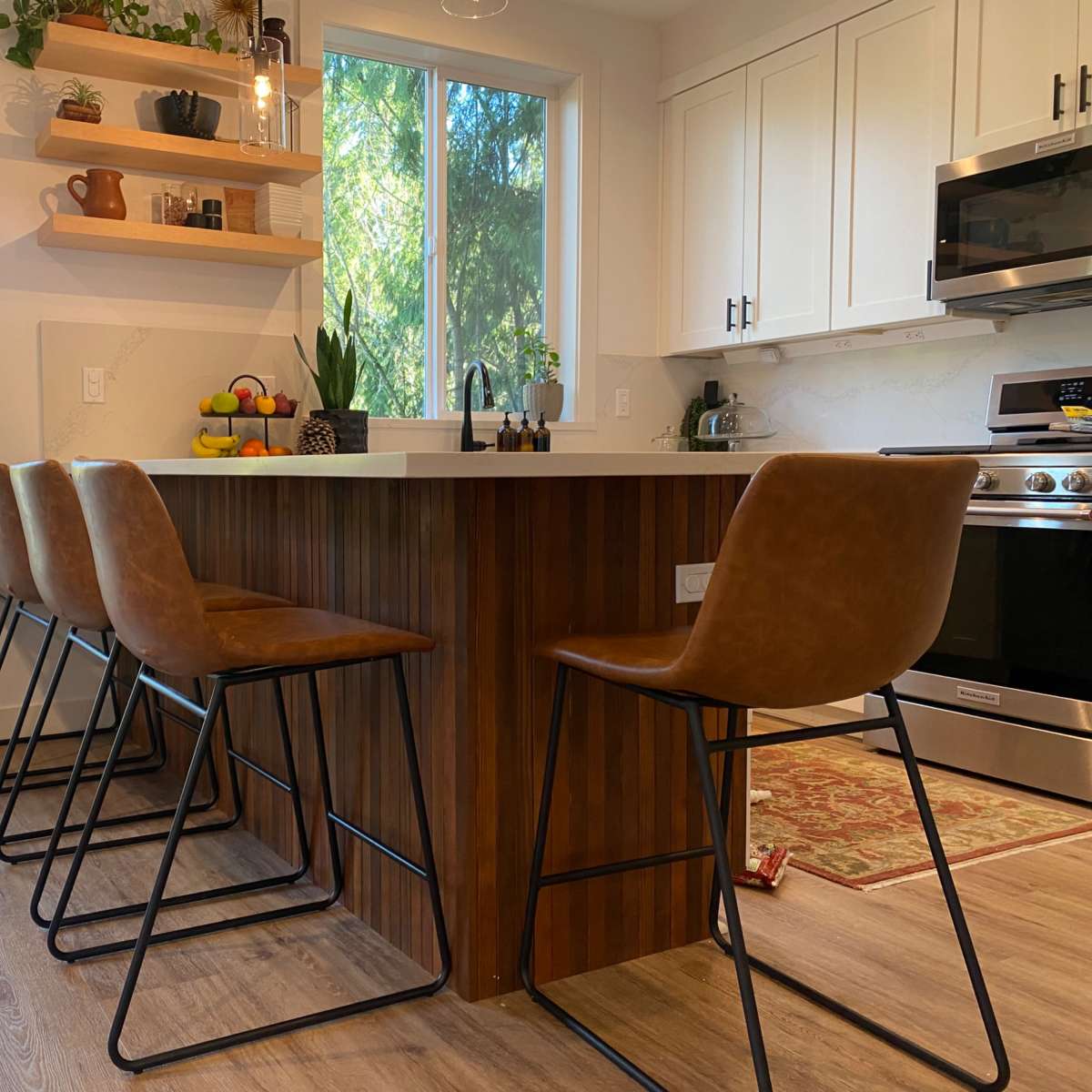 A modern kitchen with a wooden island bar and six brown leather stools, alongside a window that offers a view of greenery outside.