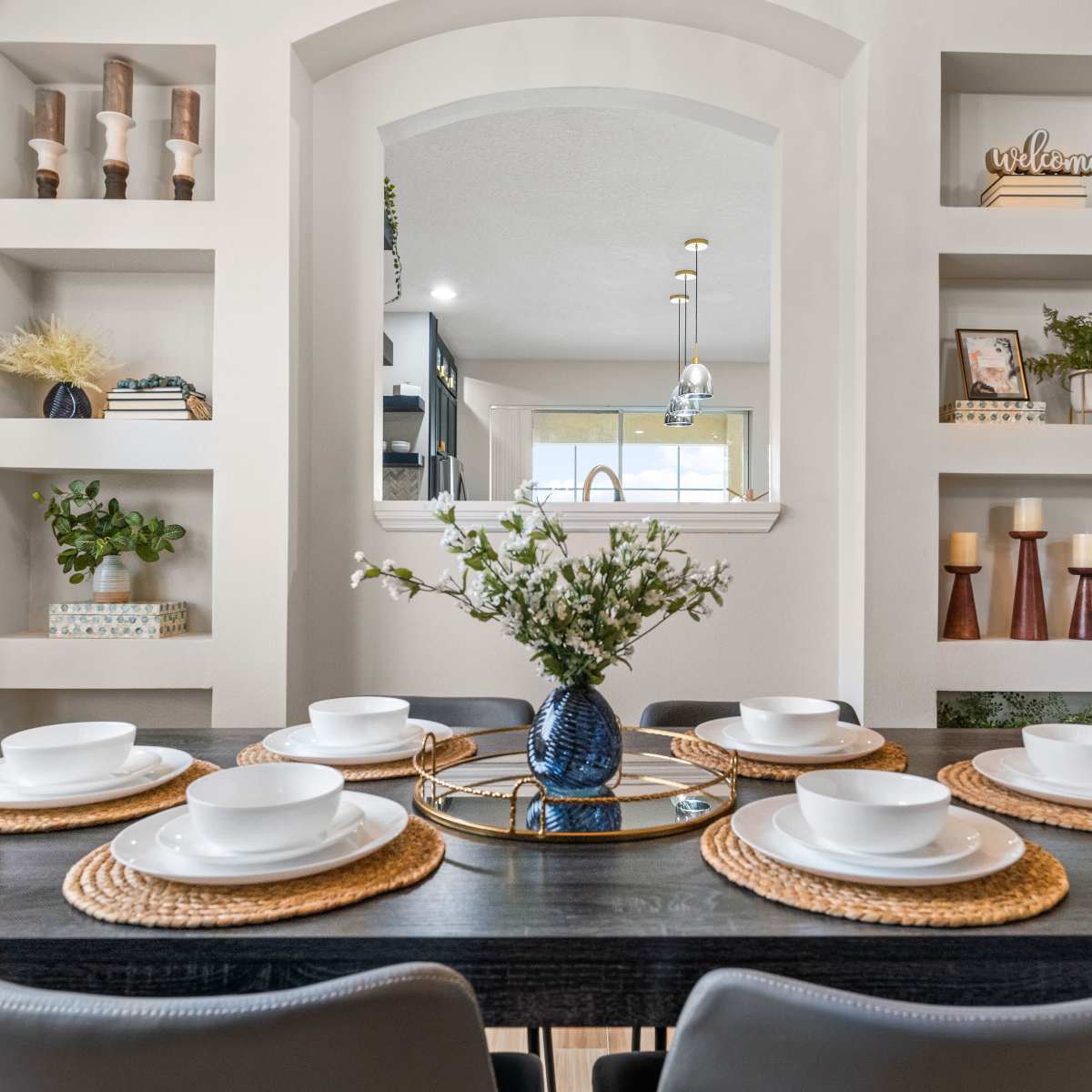 A dining table is set with six white plates on woven placemats, featuring a decorative vase of flowers at the center, surrounded by built-in shelves displaying various decorative items.