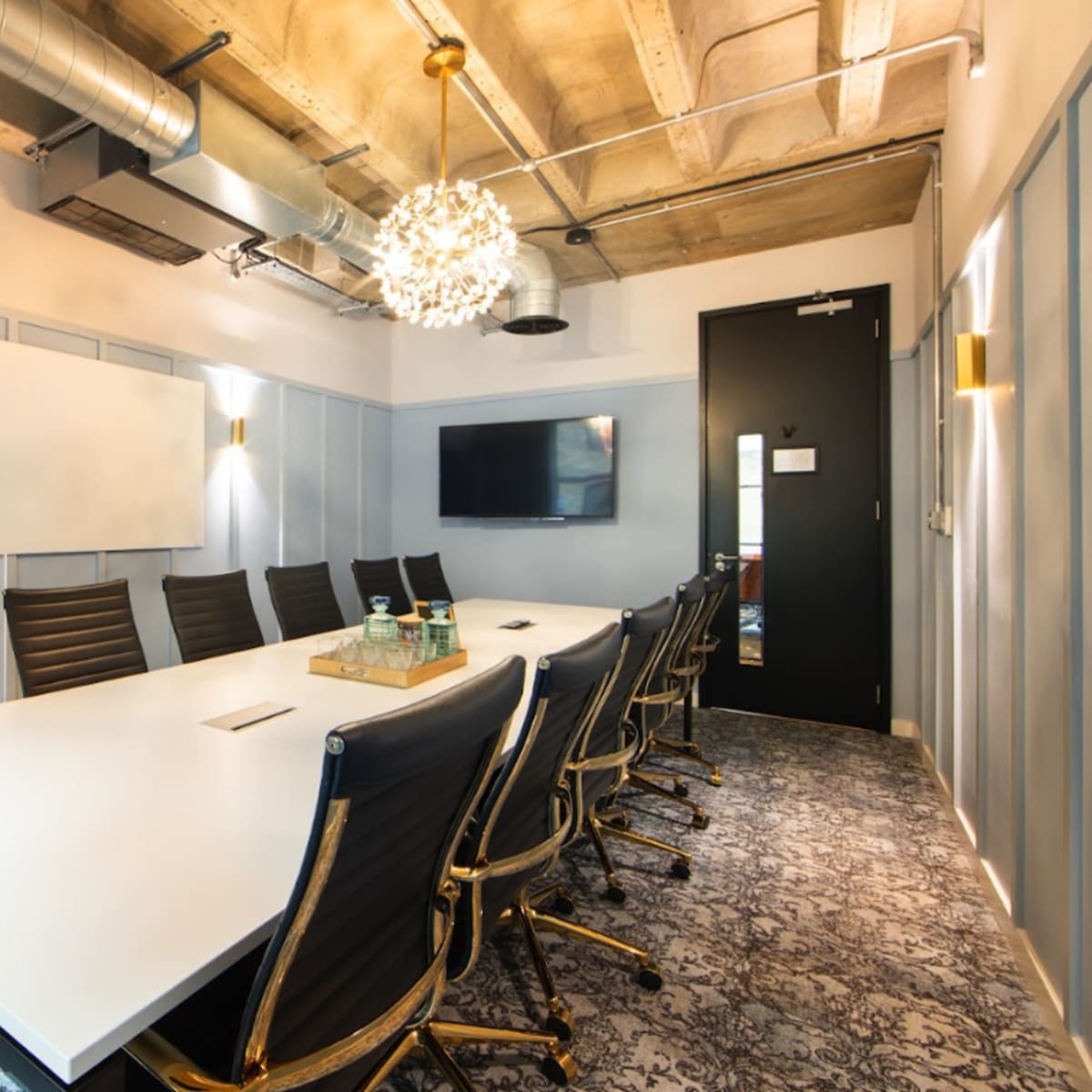 A modern conference room featuring a long rectangular table surrounded by black leather chairs, with a whiteboard and a television mounted on the wall.