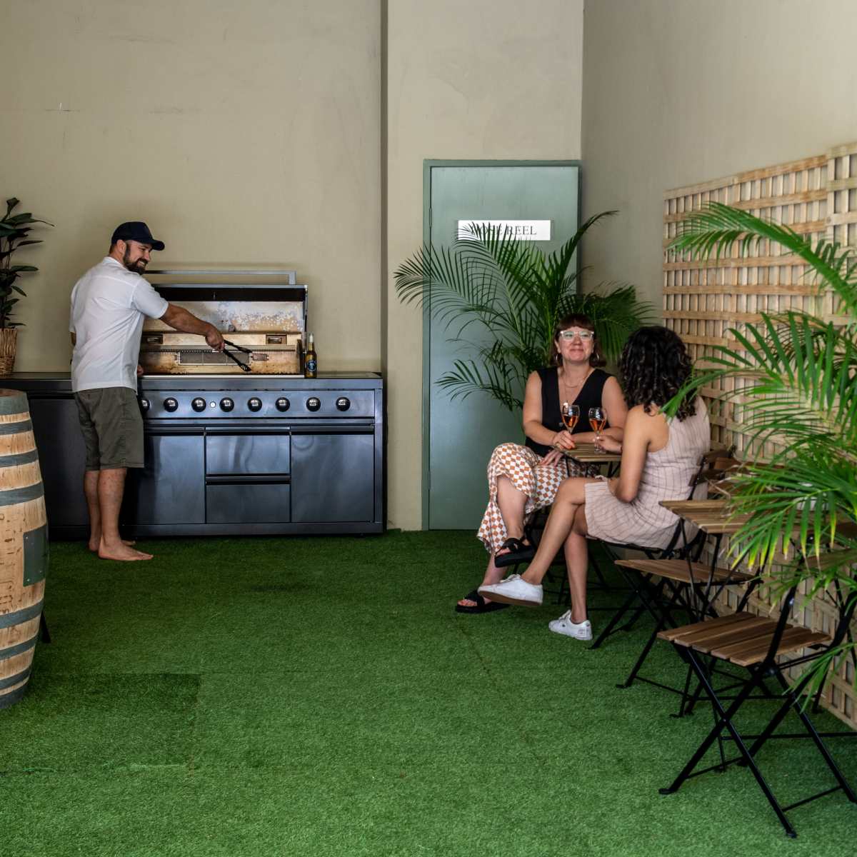 A person cooks at a grill while two women sit at a small table surrounded by potted plants in a well-lit indoor space.