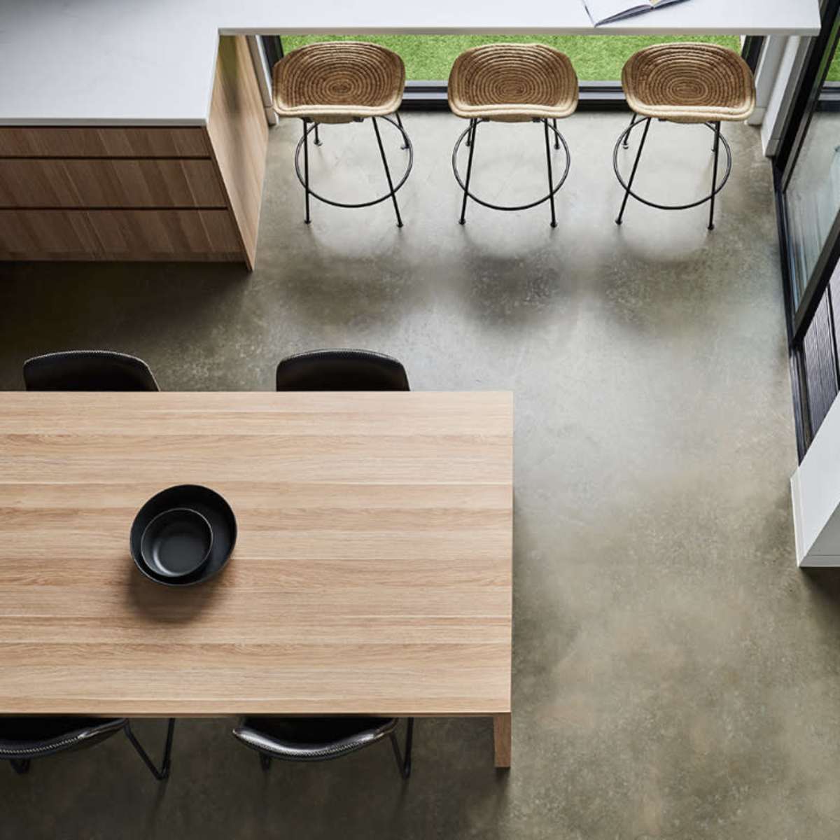 A modern kitchen and dining area with a wooden table, black chairs, and woven barstools arranged near a large window.