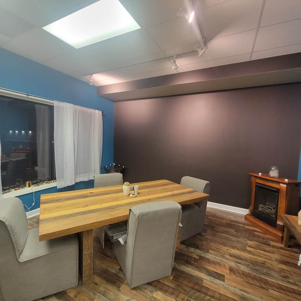 A modern room with a wooden dining table surrounded by upholstered chairs, a cozy fireplace in the corner, and a large window with sheer curtains.