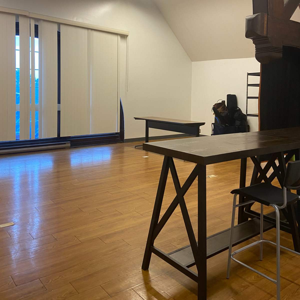 A spacious room with wooden floors, featuring a large window covered by vertical blinds, two tables, and a ladder against the wall.