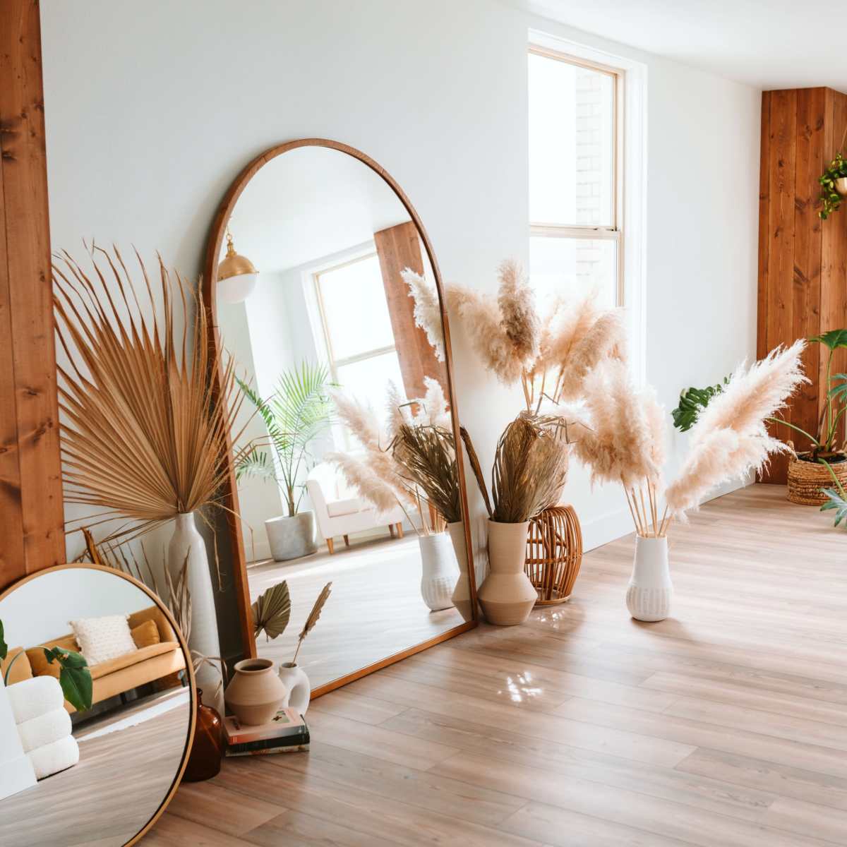 A bright room features large mirrors, decorative plants, and pampas grass arranged near wooden walls and large windows.