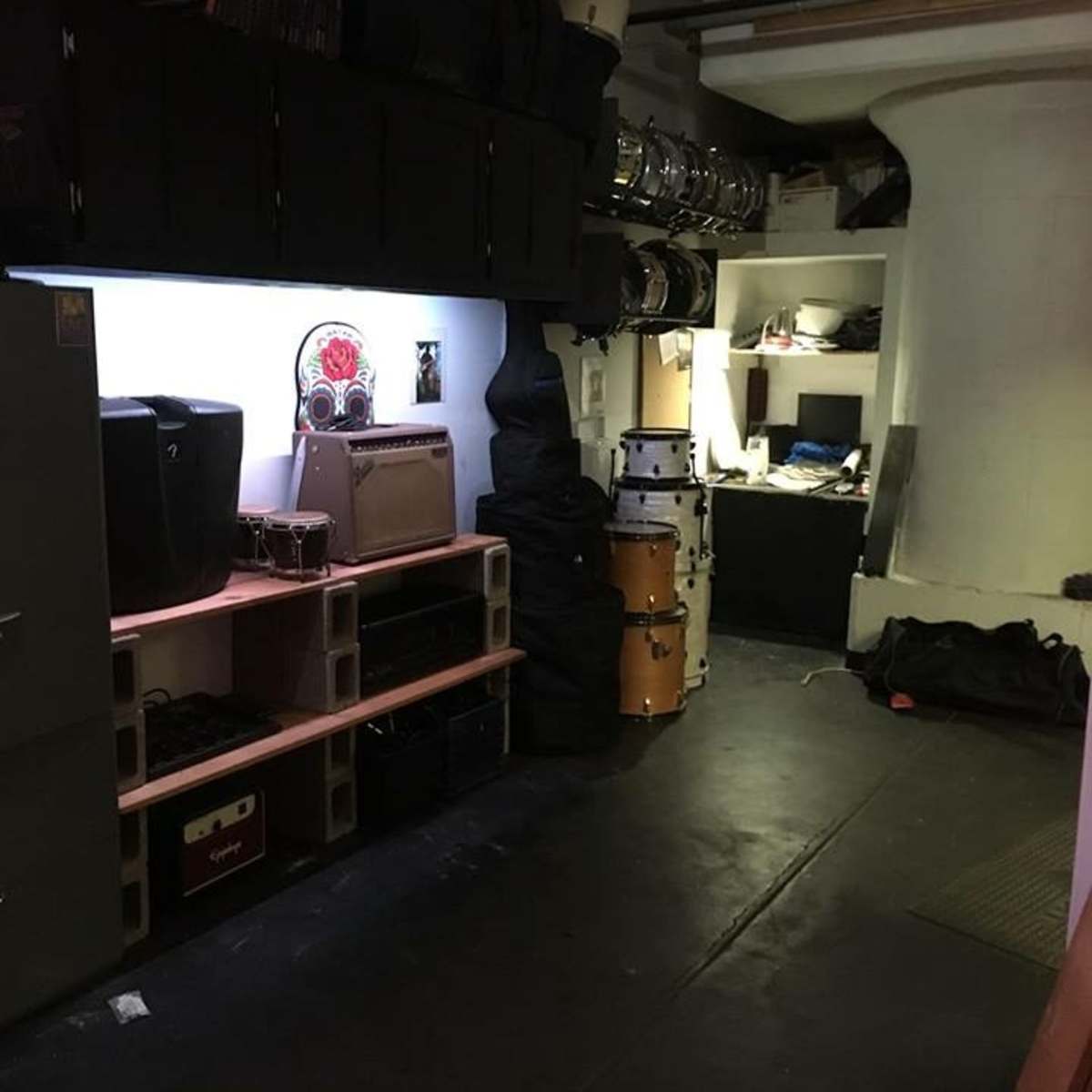 A dimly lit storage room featuring music equipment, including amplifiers and drum kits, organized on shelves and stacked on the floor.