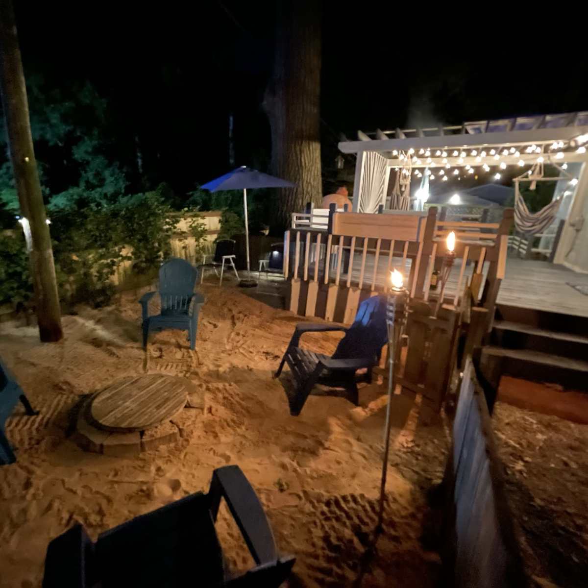 A sandy outdoor area at night with several blue chairs arranged around a fire pit, adjacent to a deck with string lights and a table under an umbrella.