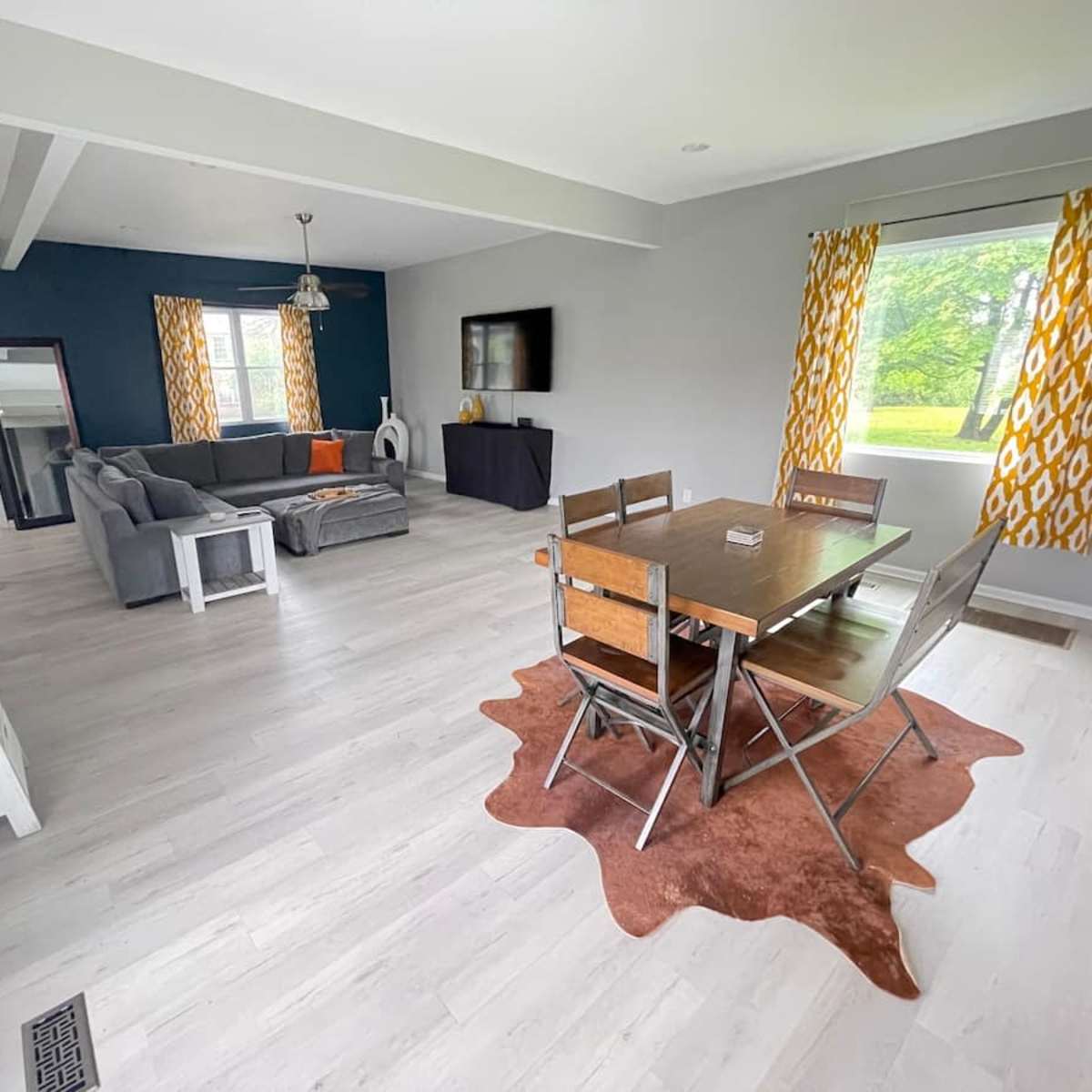 A modern, open-concept living and dining area featuring a gray sofa, a wooden dining table with metal chairs, and large windows with patterned yellow curtains.