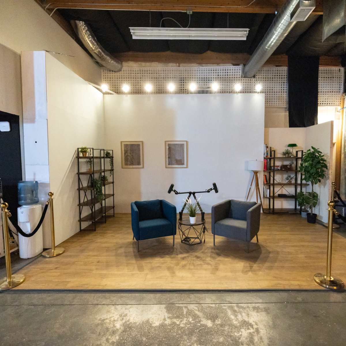 A minimalist studio setup featuring two chairs, a small table, and decorative plants, all arranged on a wooden floor with a backdrop of white walls and ambient lighting.