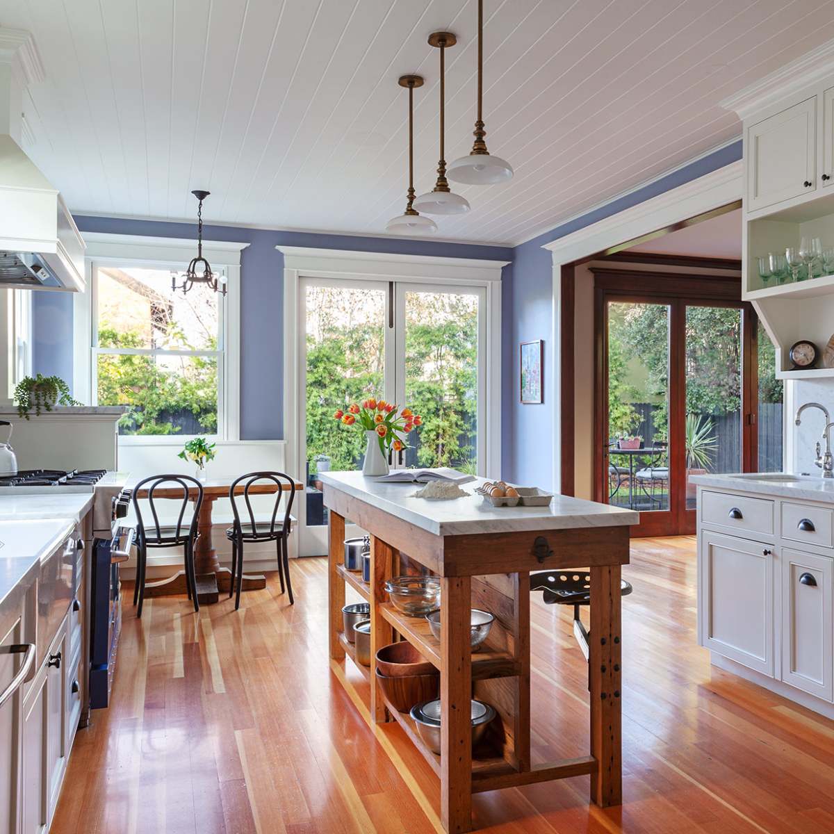 The kitchen features white cabinets, a large wooden island, and large windows overlooking greenery.