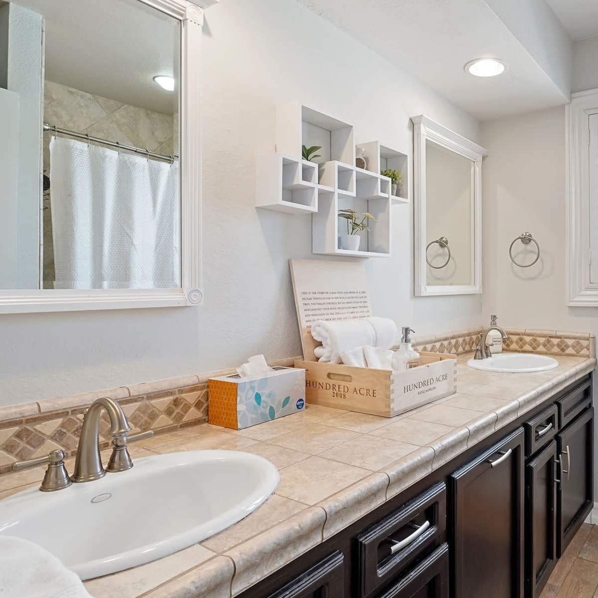 A well-organized bathroom features dual sinks with a tiled countertop, white cabinetry, and natural light filtering through a window with shutters.