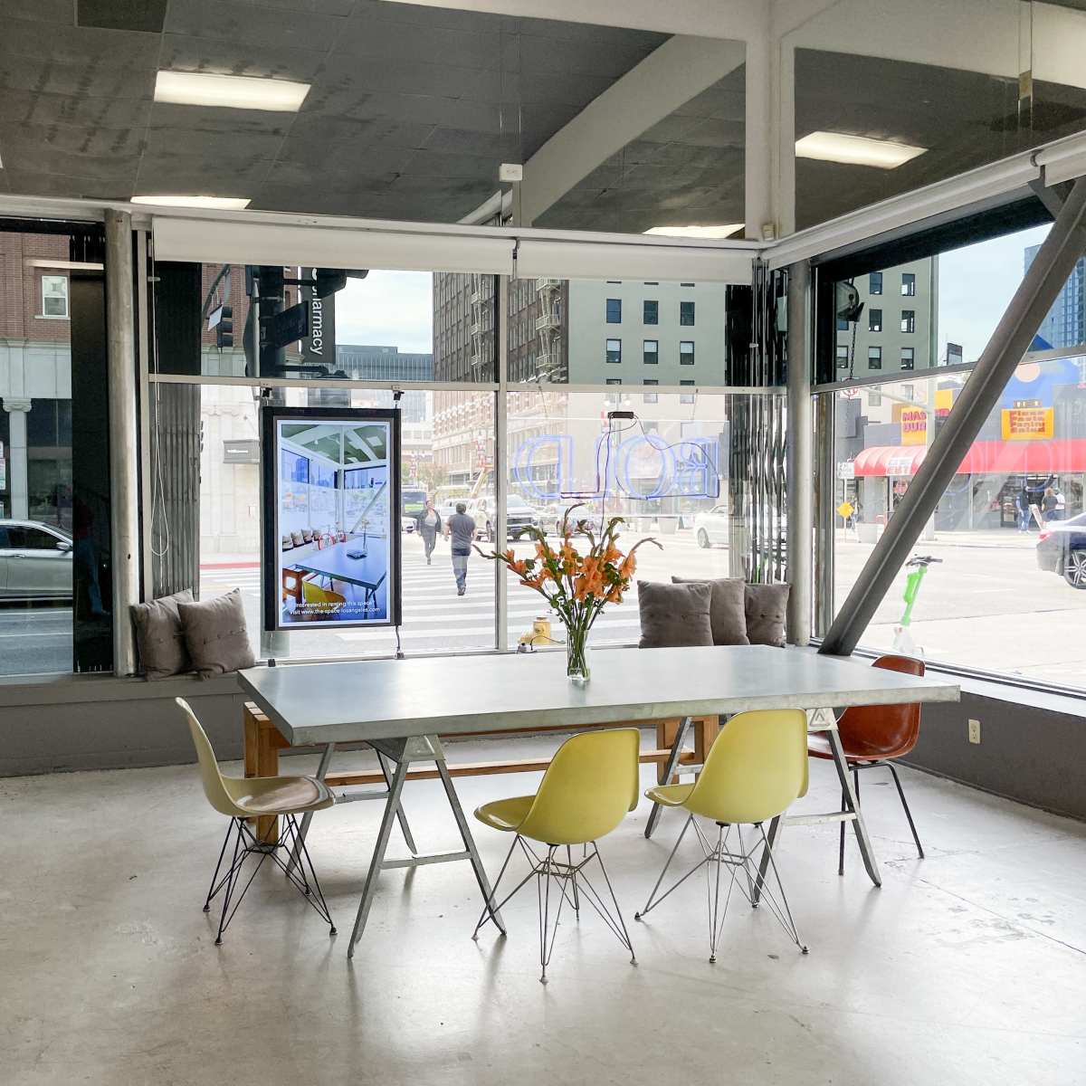 A spacious indoor setting features a large table surrounded by several chairs, with large windows offering a view of a city street.