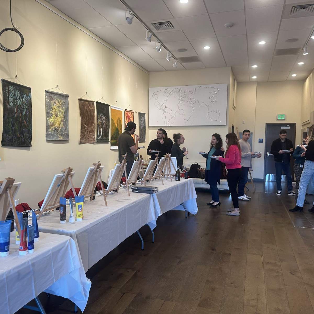 A group of people is gathered in a studio environment with easels set up on tables, canvases ready for painting, and various art supplies displayed.