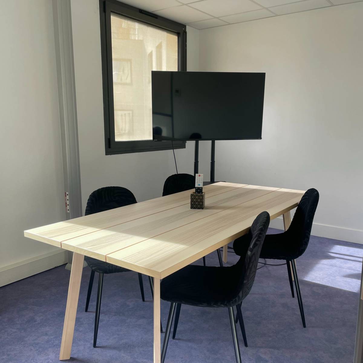 A simple meeting room with a rectangular wooden table and four black chairs, accompanied by a wall-mounted television.