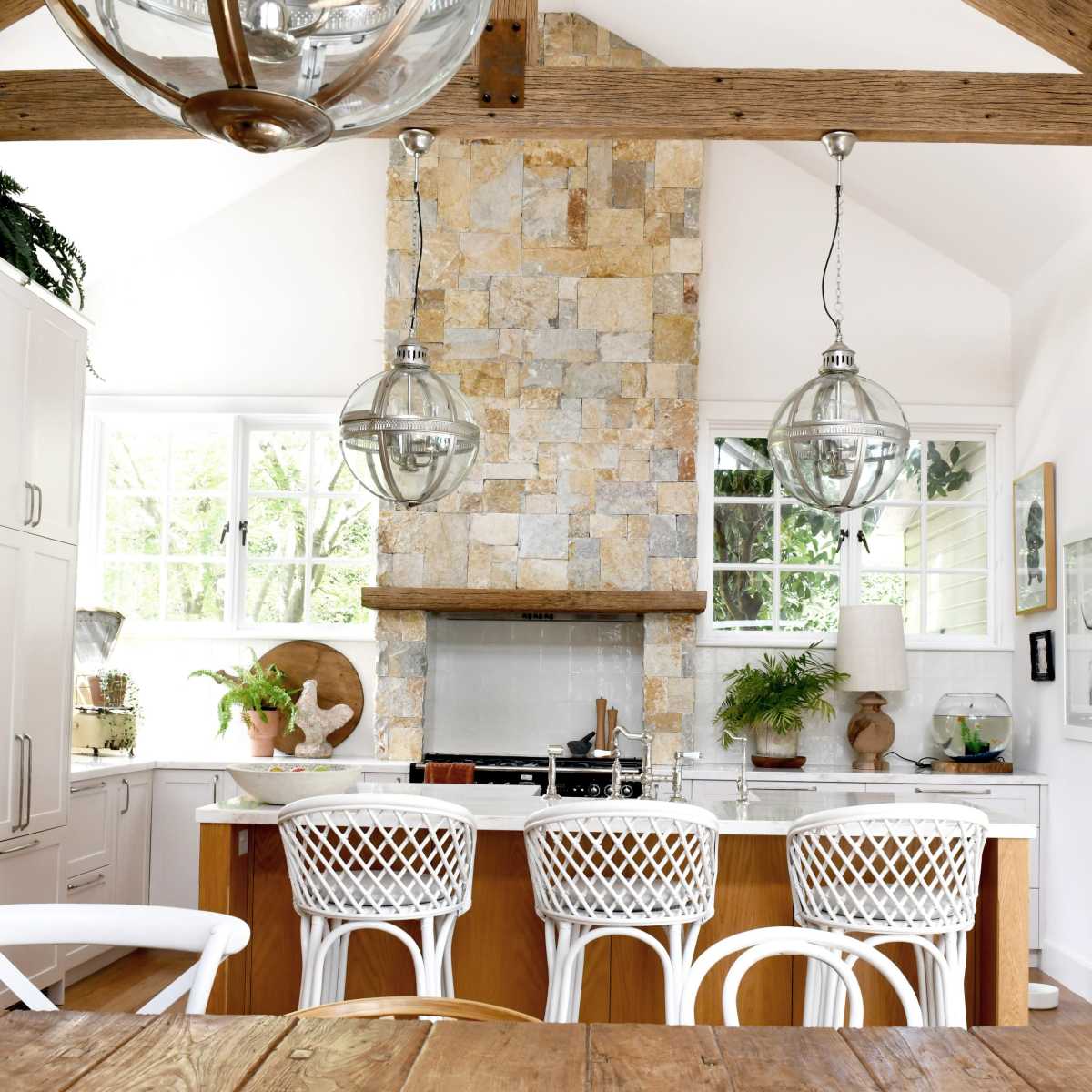 A modern kitchen with a stone accent wall, white cabinetry, and four white barstools arranged around a wooden island.