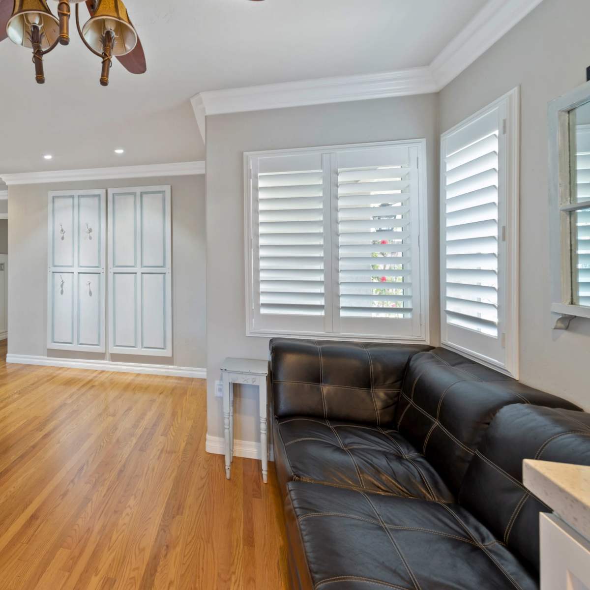 A living room with hardwood floors, a black leather couch, a white vintage-style hutch, and shuttered windows.