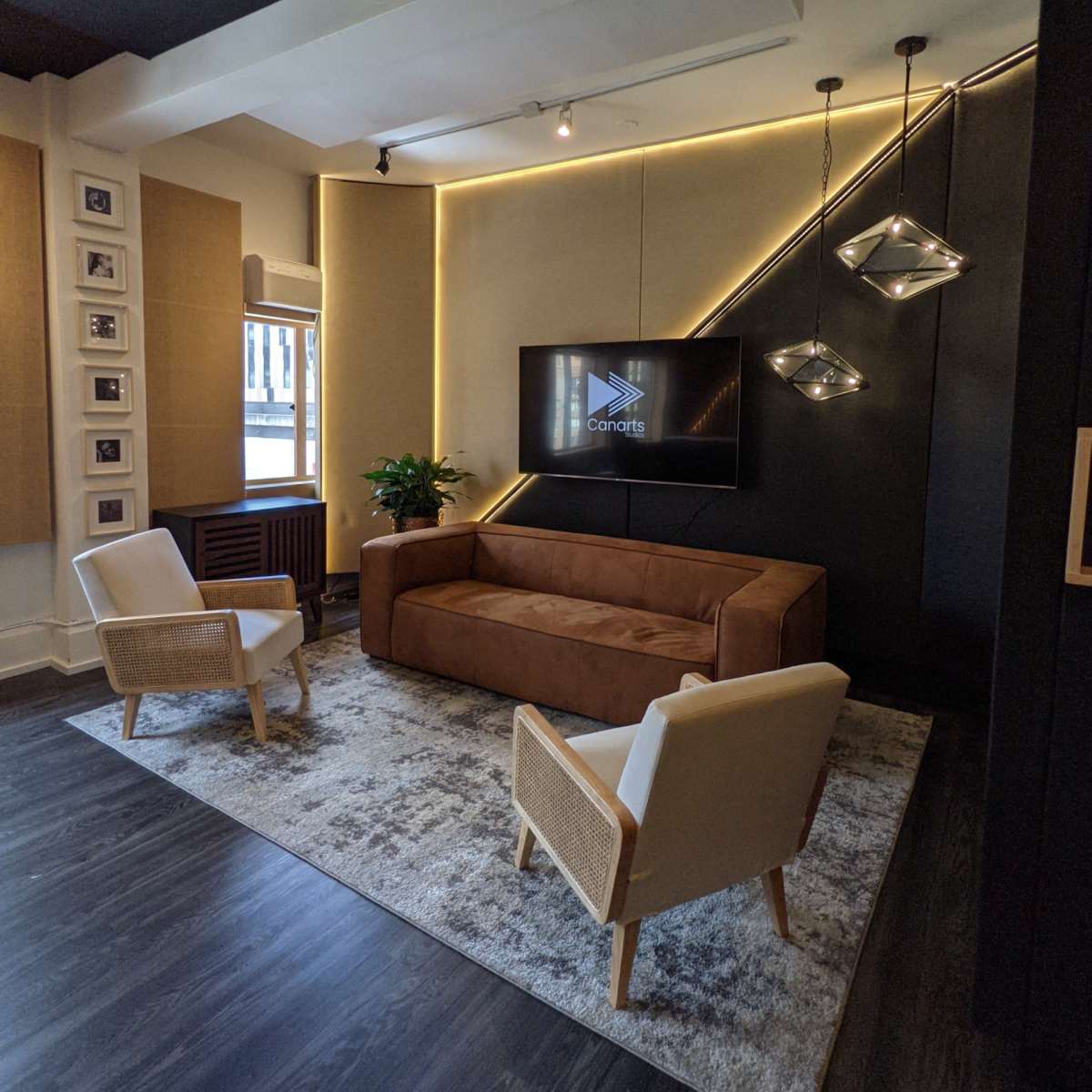 A modern living space featuring a brown couch, two white armchairs, a large television mounted on a black wall, and decorative lighting overhead.