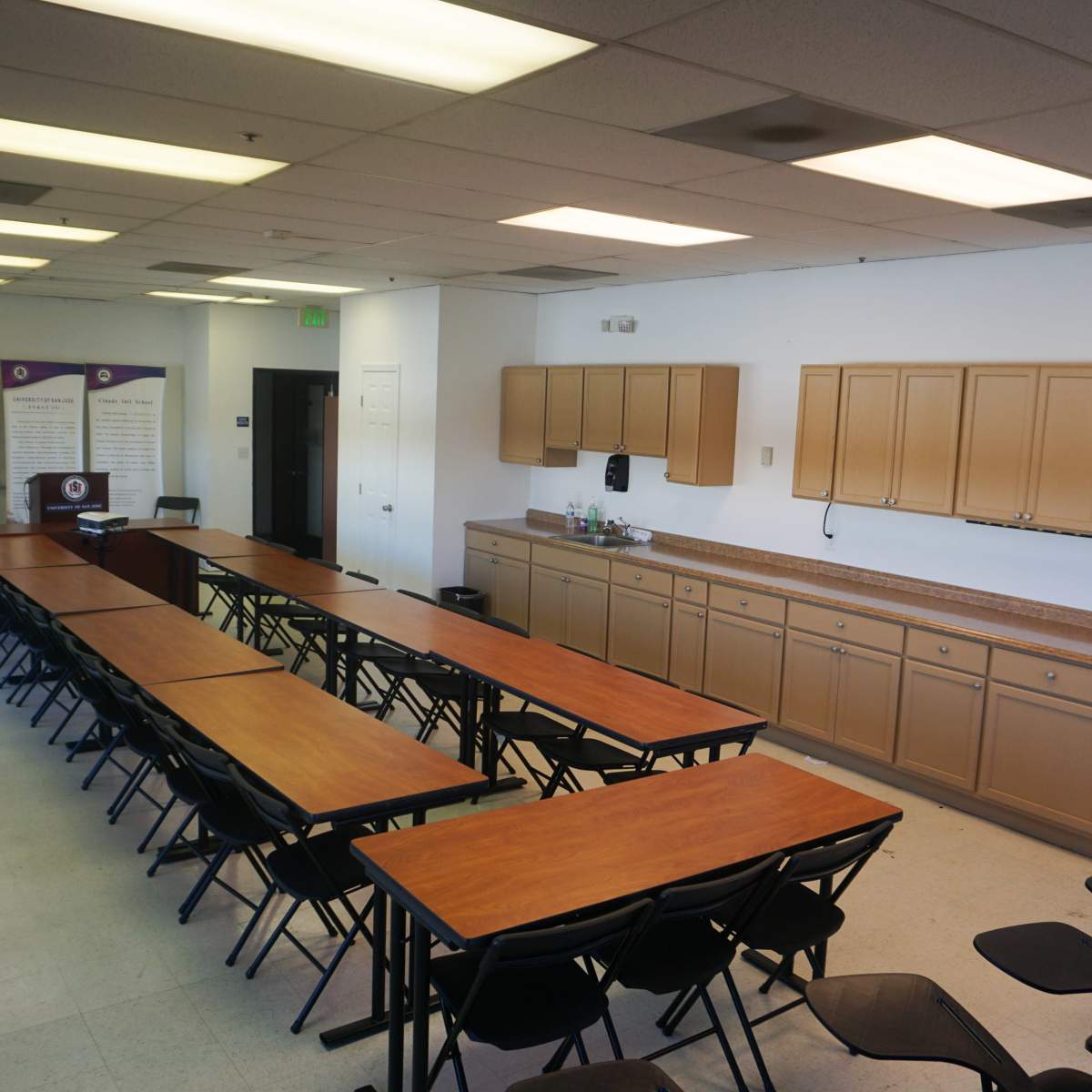 A spacious meeting room with several rows of rectangular tables and chairs arranged to facilitate presentations, along with a kitchen area featuring cabinets and a countertop.