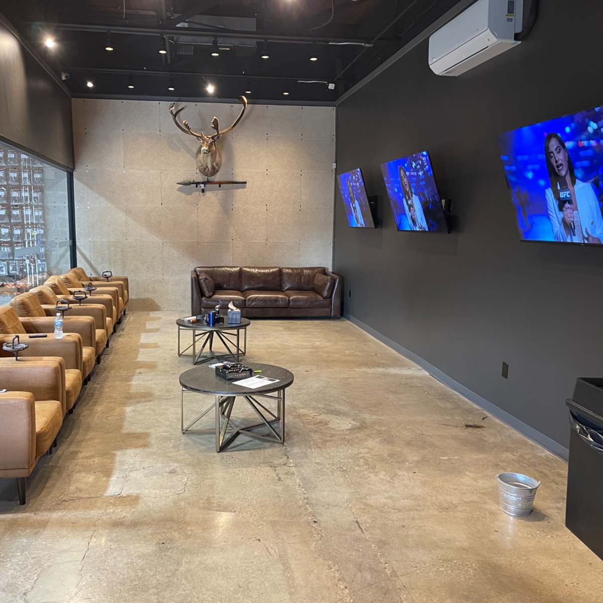 A modern lounge area with a brown leather couch, several tan chairs, two mounted TVs displaying a news program, and a decorative antler sculpture on the wall.