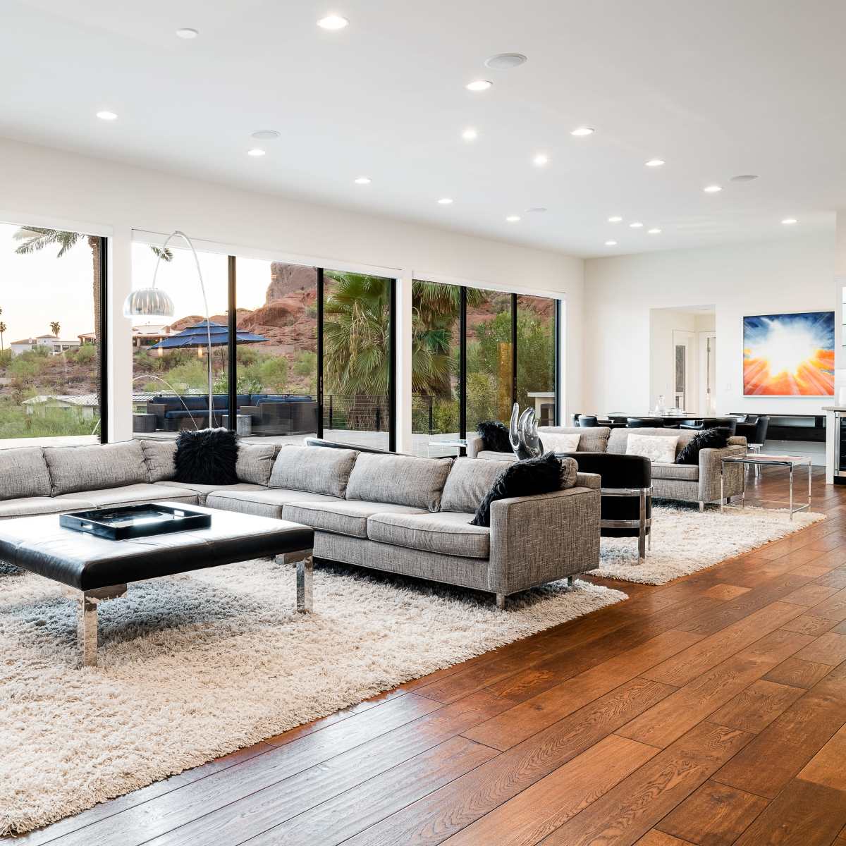 The living room features a large gray sectional sofa, a black coffee table, and expansive windows that provide views of palm trees and a natural landscape.