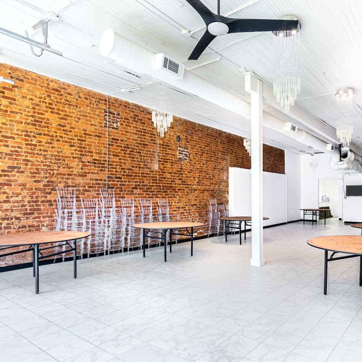 An empty event space featuring several round tables surrounded by transparent chairs, with exposed brick walls and a white ceiling.