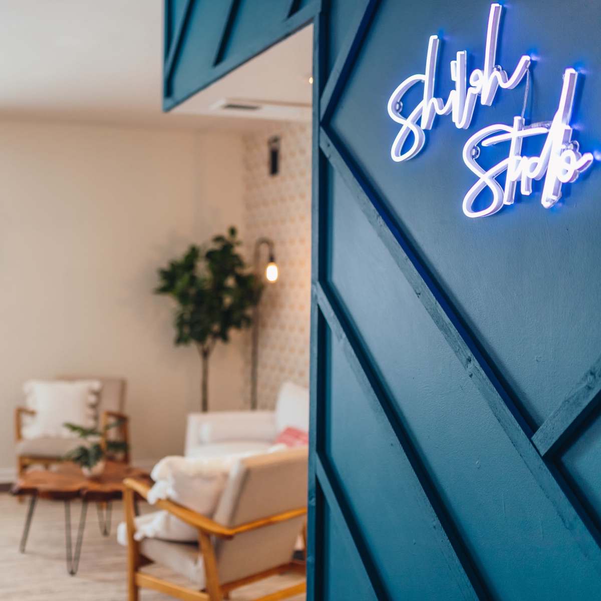 A brightly lit room with a blue wall featuring a neon sign that reads "Shulh Studio," and two chairs positioned around a small coffee table.