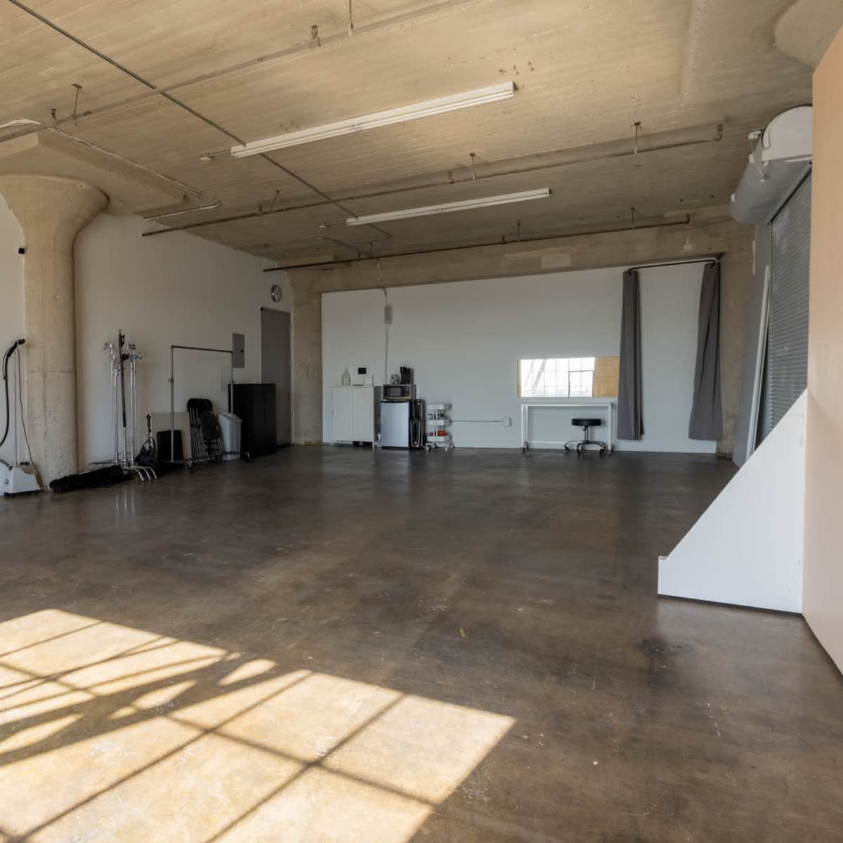 A spacious, empty room with a concrete floor, large windows, and minimal furnishings, including a small kitchenette and some workout equipment.