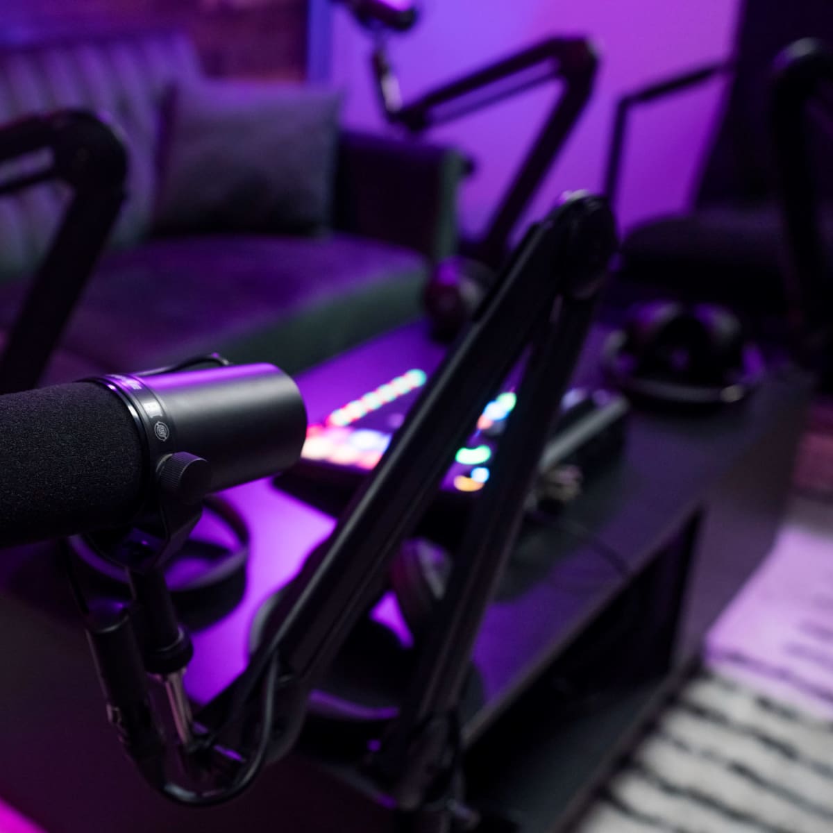 A black microphone is positioned on a stand in a podcasting studio, with a green couch and colorful soundboard visible in the background.