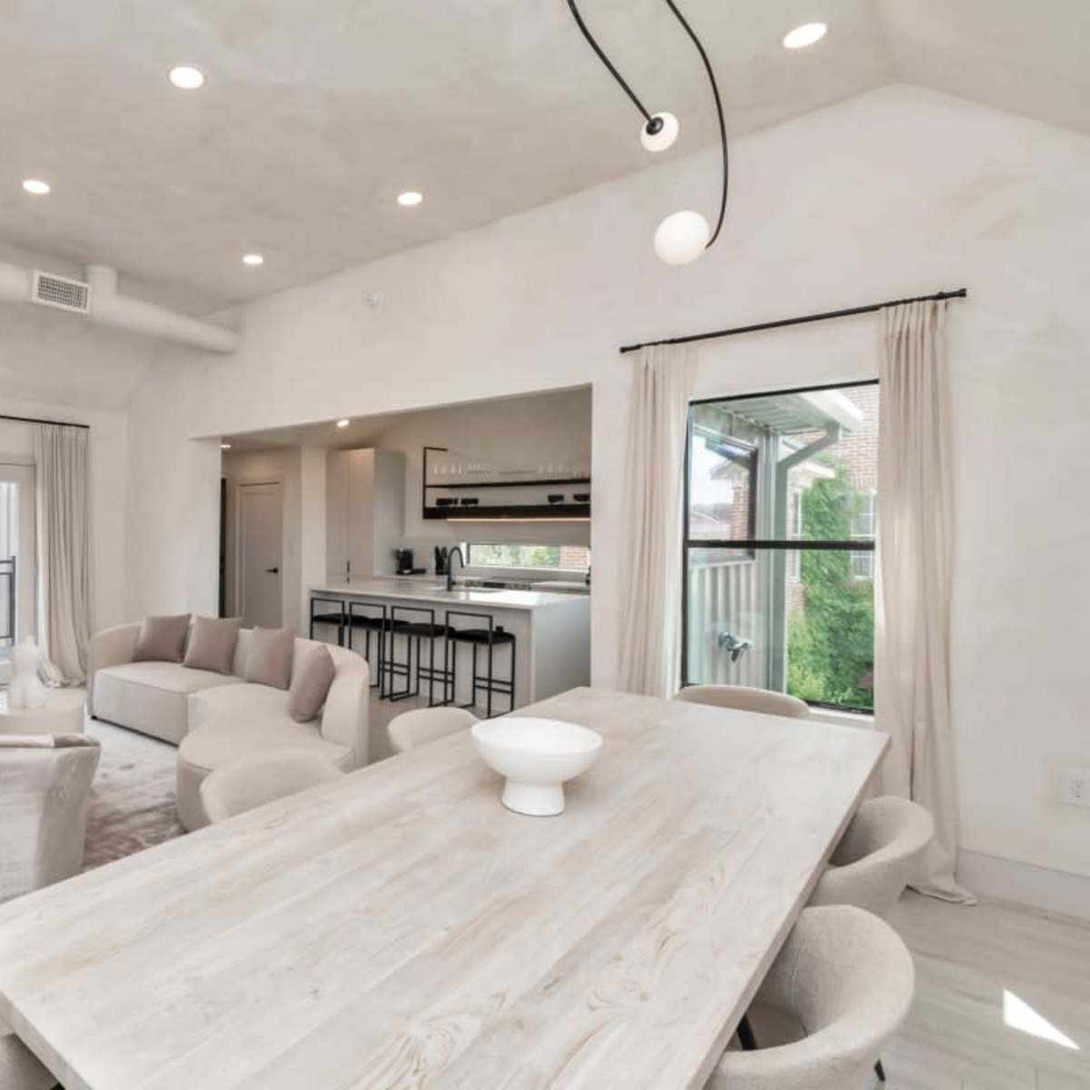 A modern, open-concept living space with a light-colored interior, featuring a dining table, comfortable seating, and large windows leading to a balcony.