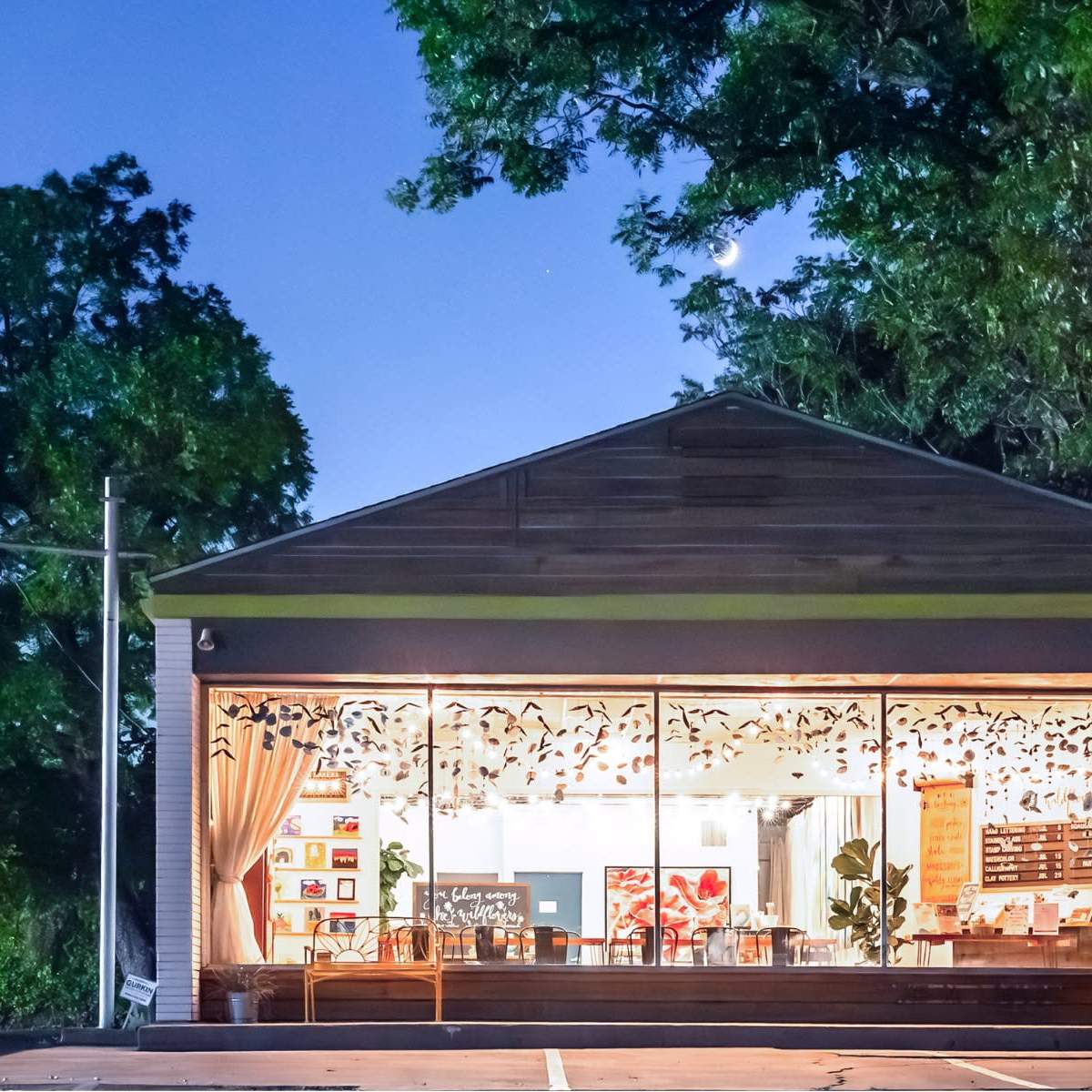 A brightly lit café with large front windows showcasing interior seating is set against a backdrop of trees at dusk.