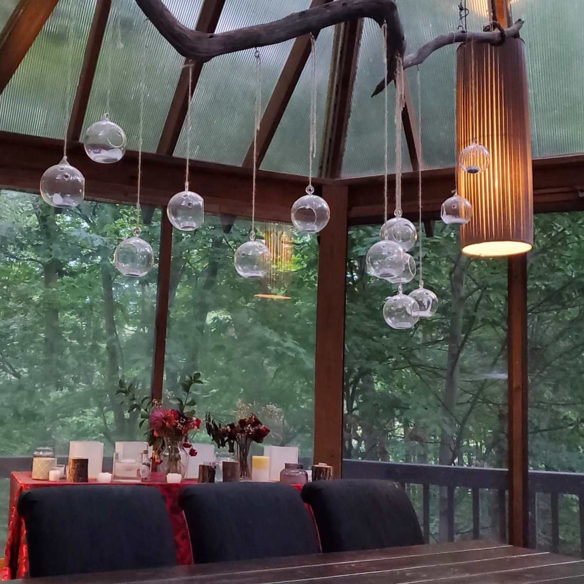 A rustic dining area with a wooden table surrounded by chairs, illuminated by hanging glass globes and cylindrical light fixtures, with greenery visible through the glass walls.