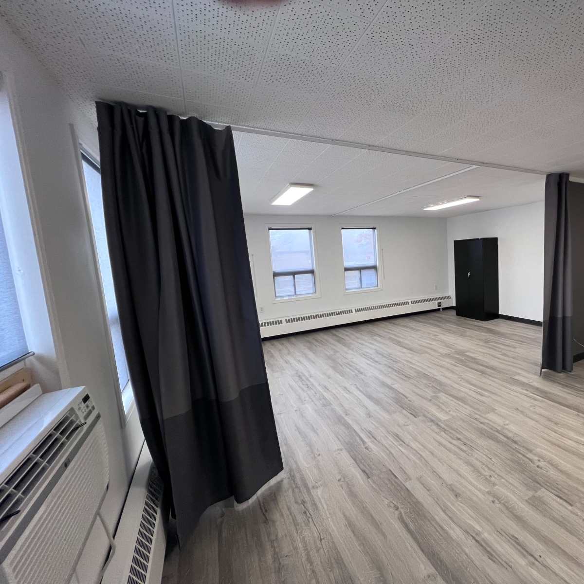 A spacious, empty room with gray curtains, a standing desk, and large windows that allow natural light.