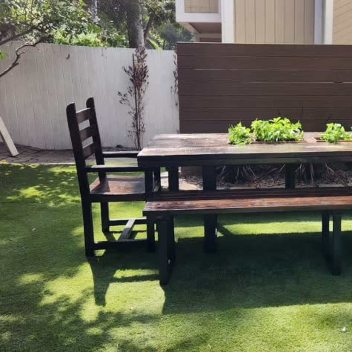 A wooden picnic table and benches are positioned on a grassy area surrounded by a fence and a wall with plants in the background.