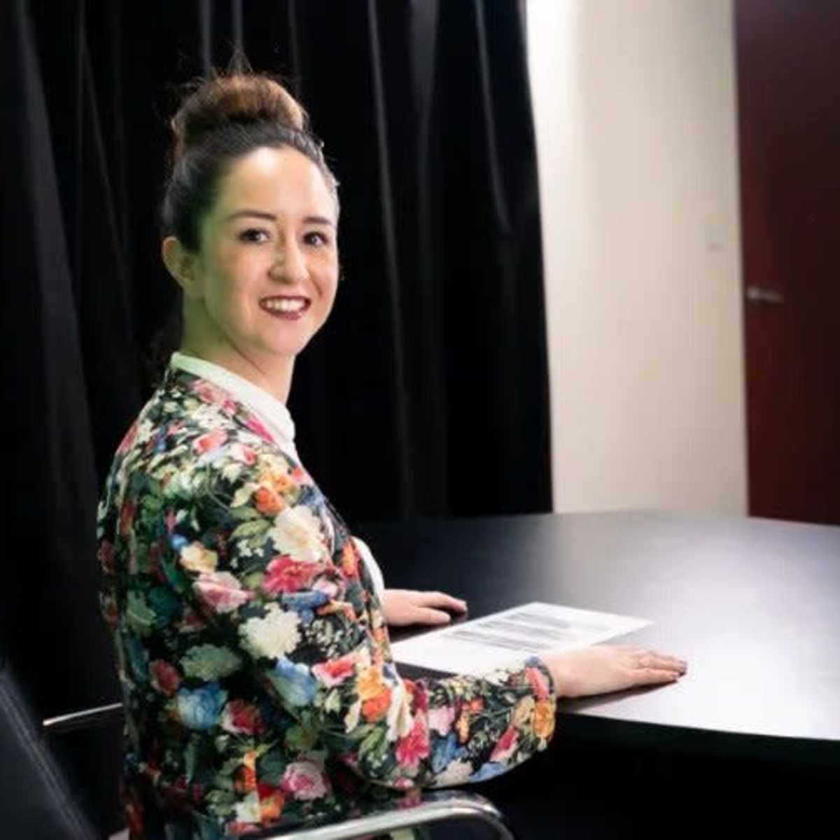 A woman sits at a black table in a studio setting, smiling while looking at the camera, with a presentation displayed on a screen behind her.