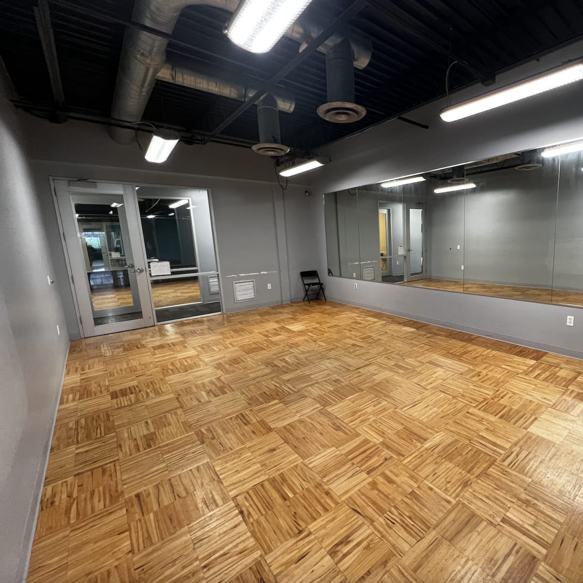A spacious room with wooden flooring, mirrored walls, and a few black folding chairs arranged along one side.