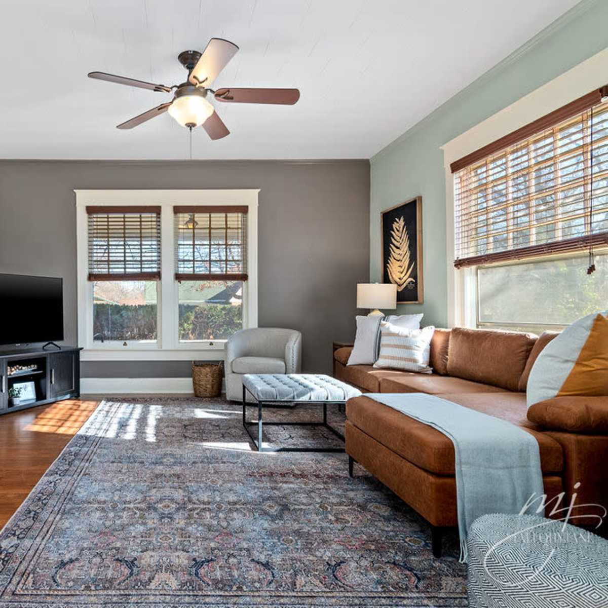 A living room featuring a brown sofa, a television on a black stand, and a patterned rug on wooden floors, with natural light coming through the windows.