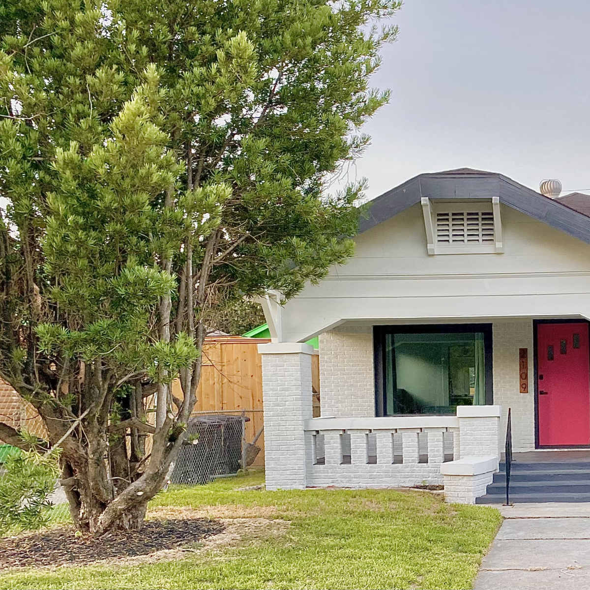 A single-story, white-painted house with a pink front door, situated on a grassy lot with a large tree in the front yard.