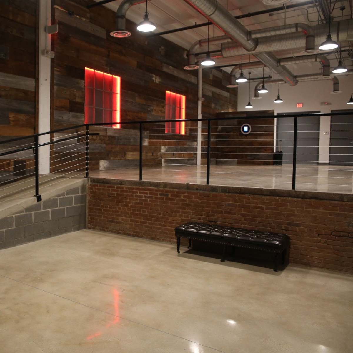 A spacious interior area with a wooden wall, exposed pipes, a metal railing, and a flat bench along a concrete floor.