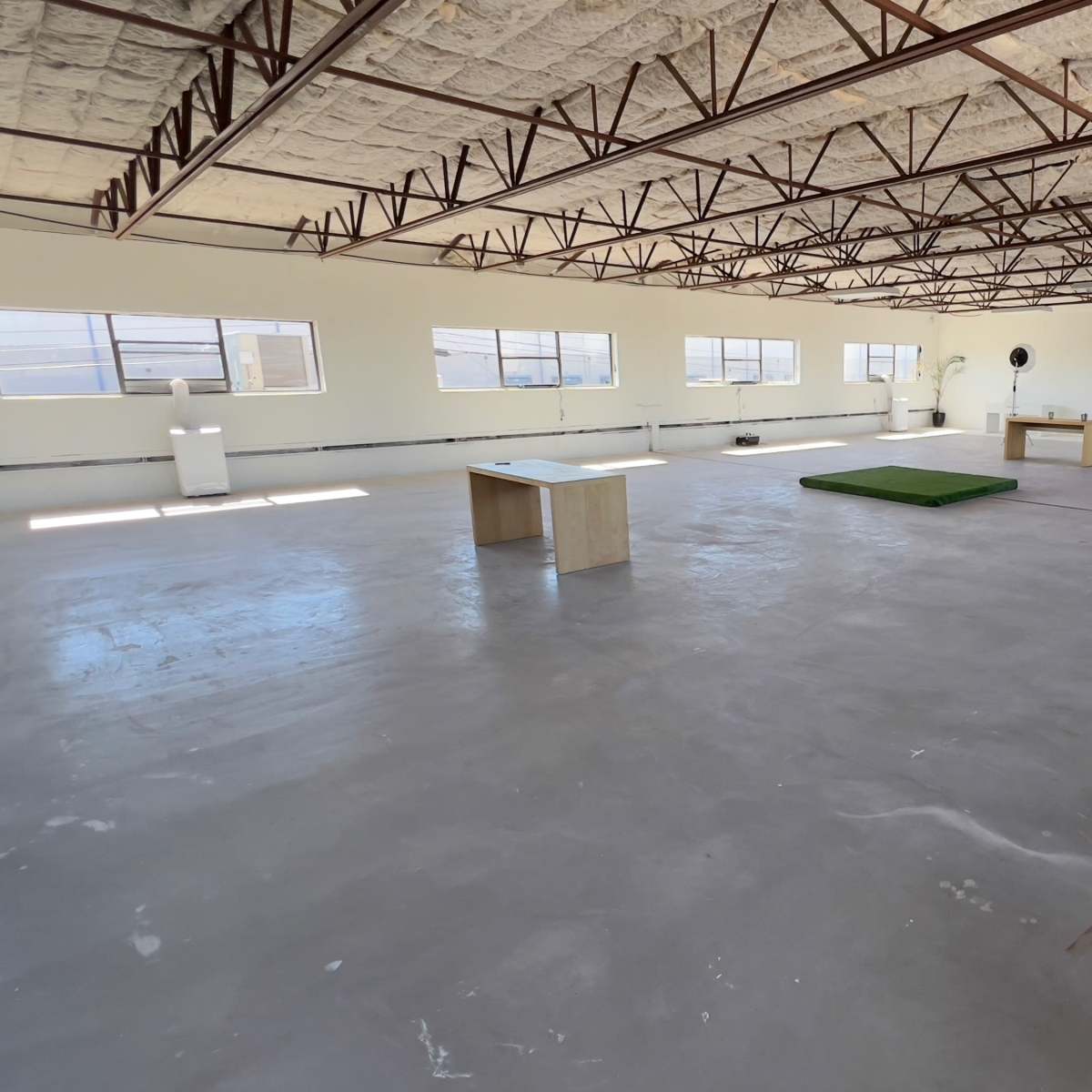 A large, empty room with a concrete floor, several windows, and minimal furnishings including a table and a green mat.