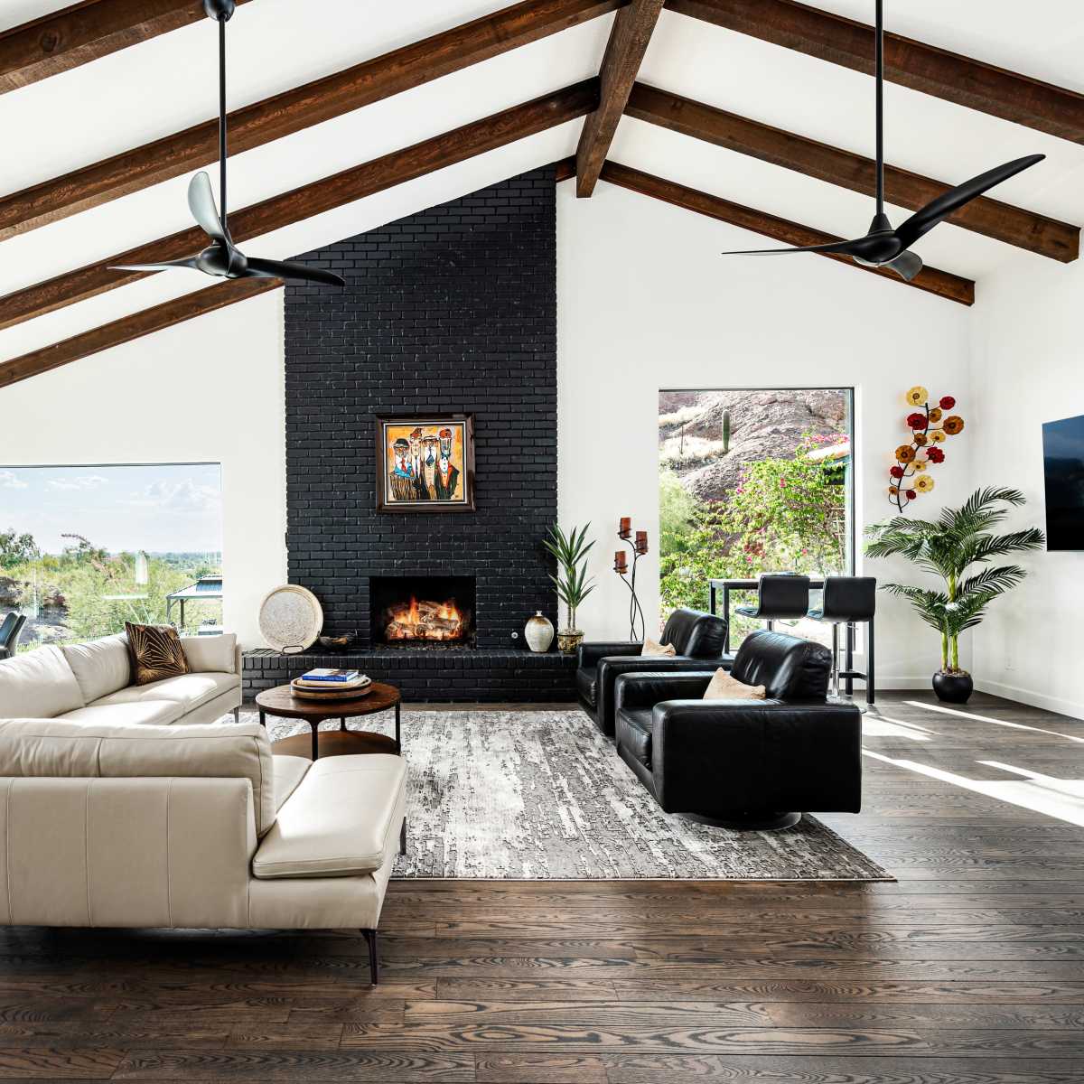 The living room features a white sofa, black accent chairs, a large stone fireplace, and wood beams on the ceiling, with large windows providing natural light and a view of the outdoors.