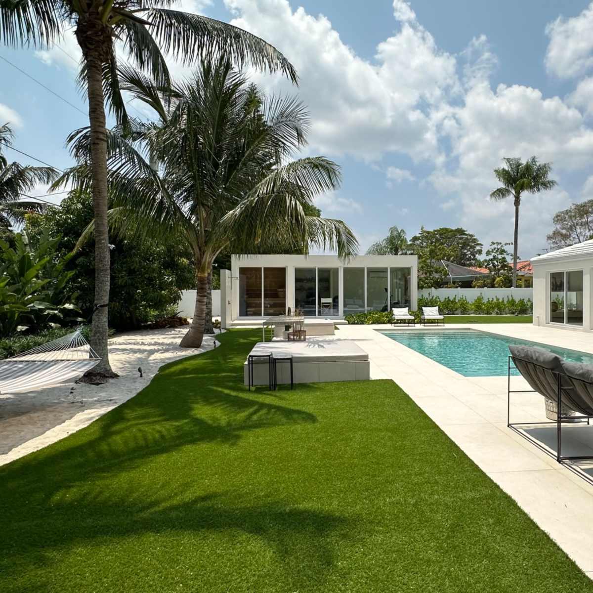 A modern outdoor space featuring a swimming pool, lounge chairs, palm trees, and two contemporary glass-walled buildings.